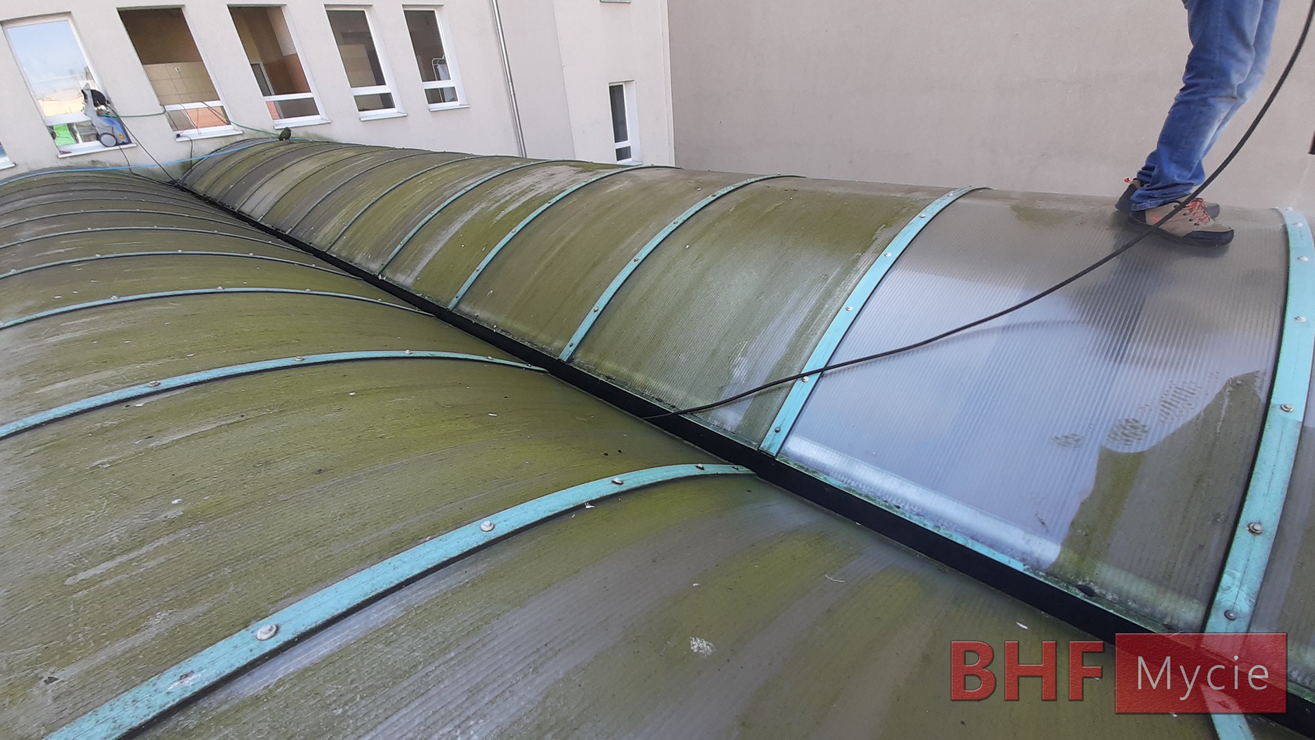Czyszczenie świetlika poliwęglanowego na dachu budynku, widoczne ślady mycia pod ciśnieniem i osoba w jeansach oraz obuwiu roboczym, w tle okna budynku.