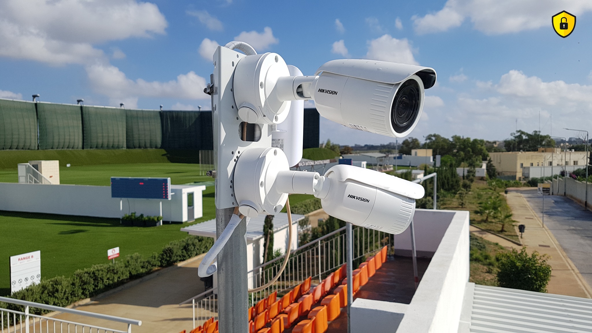 Dwie kamery monitoringu Hikvision zamontowane na metalowym słupie z widokiem na strzelnicę Malta National Shooting Ranges w słoneczny dzień.