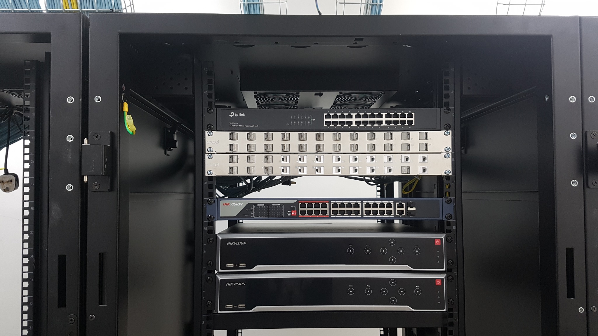 Widok z przodu szafy rack z zamontowanymi urządzeniami sieciowymi i rejestratorami wideo, widoczne switche TP-Link i Excel oraz rejestratory Hikvision, okablowanie częściowo widoczne z boku szafy.