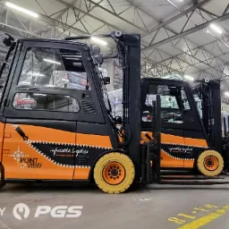 Dwa wózki widłowe elektryczne, pomalowane w barwach firmy POINT OF VIEW, stoją w magazynie wypełnionym paletami z towarem. Na wózkach widoczne logo firmy. Z przodu widoczny napis 'MADE BY PGS'.