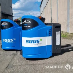 Dwa niebieskie wózki paletowe elektryczne Linde T20 z logo SUUS Logistics, stojące na betonowym placu przed budynkiem magazynowym. Widoczny kod QR i logo producenta PGS.