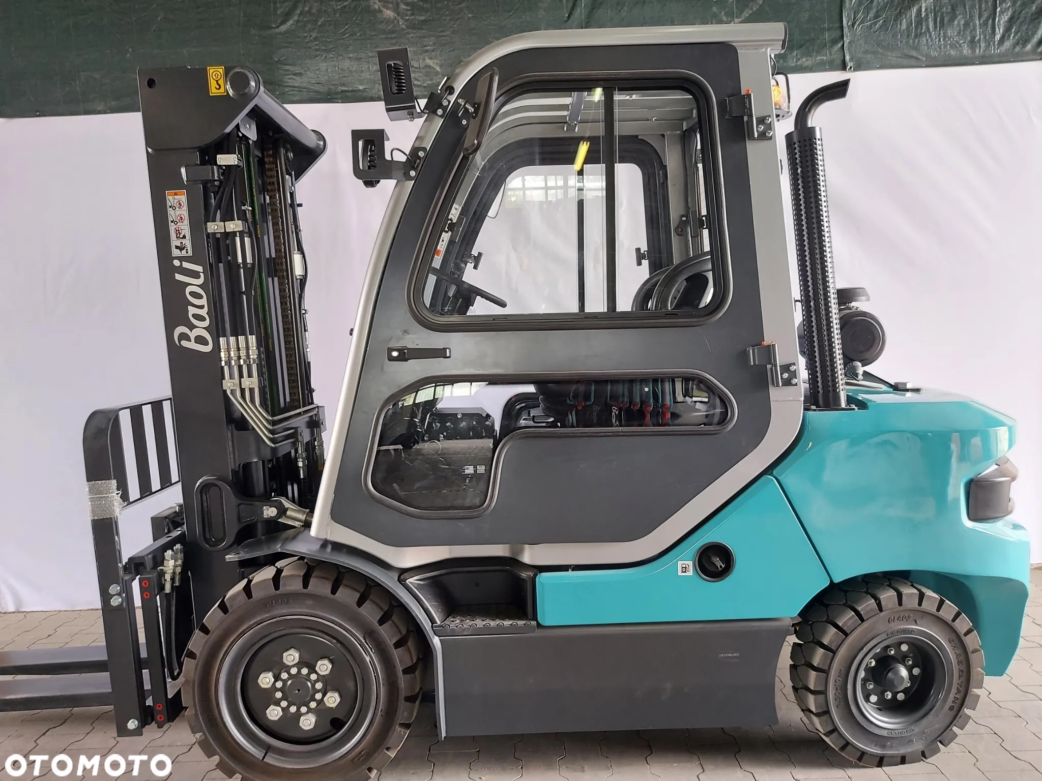 Niebiesko-szary wózek widłowy marki Baoli, widok z boku, z widoczną kabiną operatora, oponami i masztem, na tle białej ściany.