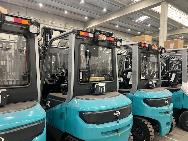 Rząd nowych, turkusowych wózków widłowych Baoli w magazynie, widoczne oświetlenie robocze i logo marki.