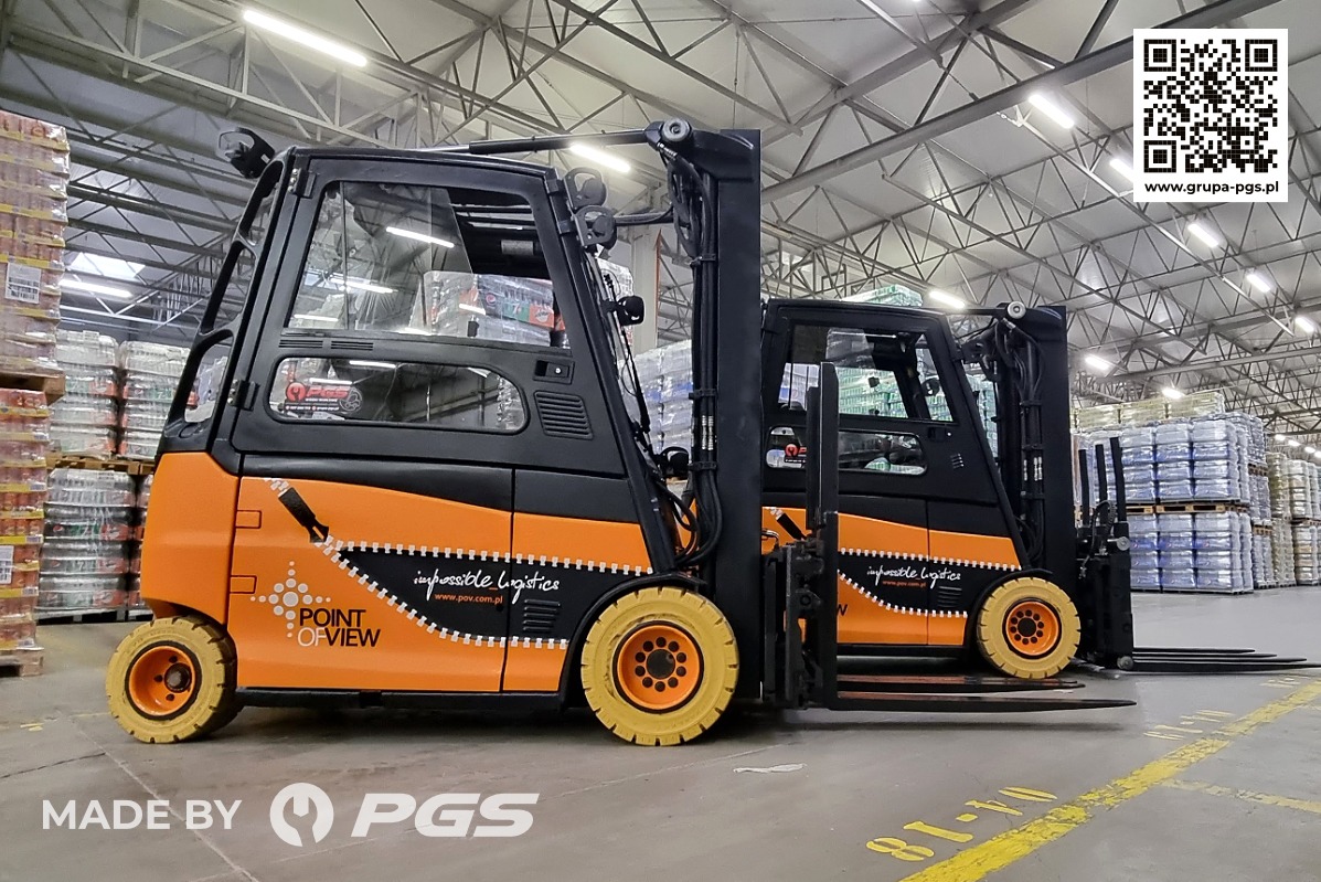 Dwa wózki widłowe elektryczne, pomalowane w barwach firmy POINT OF VIEW, stoją w magazynie wypełnionym paletami z towarem. Na wózkach widoczne logo firmy. Z przodu widoczny napis 'MADE BY PGS'.