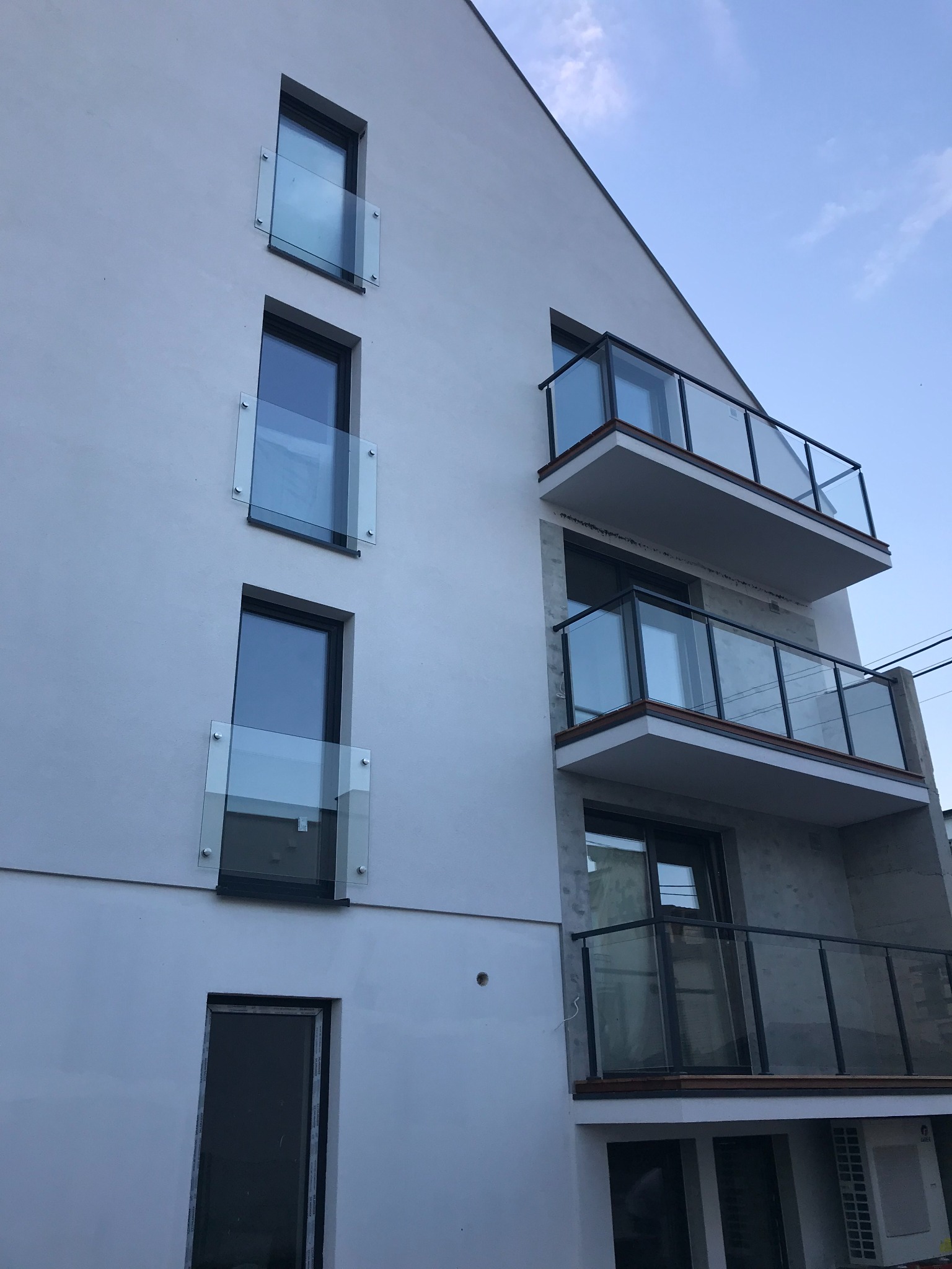 Nowoczesny budynek z białymi ścianami i balkonami z czarnymi ramami oraz szklanymi panelami, widok z dołu na błękitne niebo.