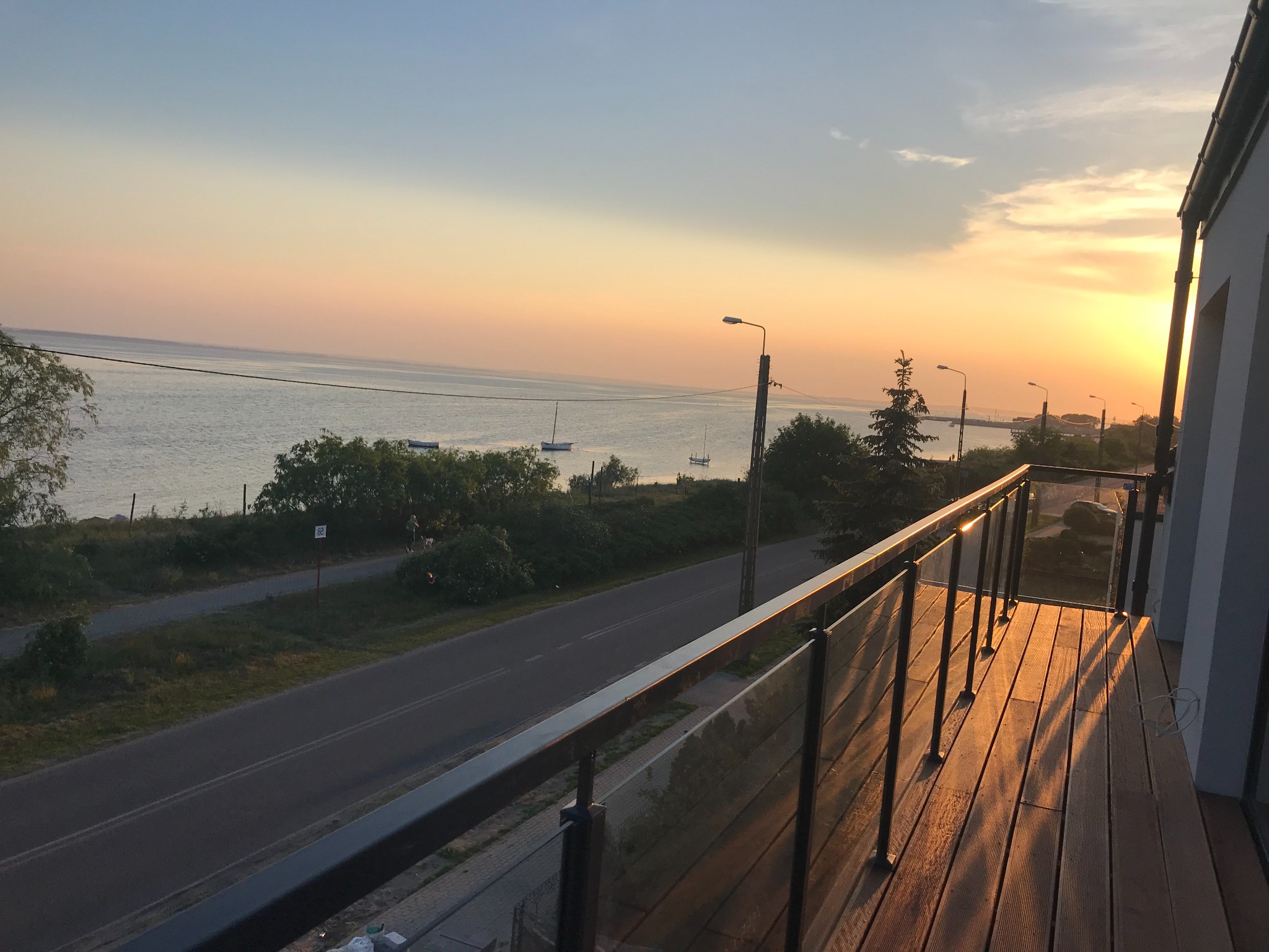 Balkon z drewnianą podłogą i szklaną balustradą, widok na morze o zachodzie słońca, ulica wzdłuż brzegu.