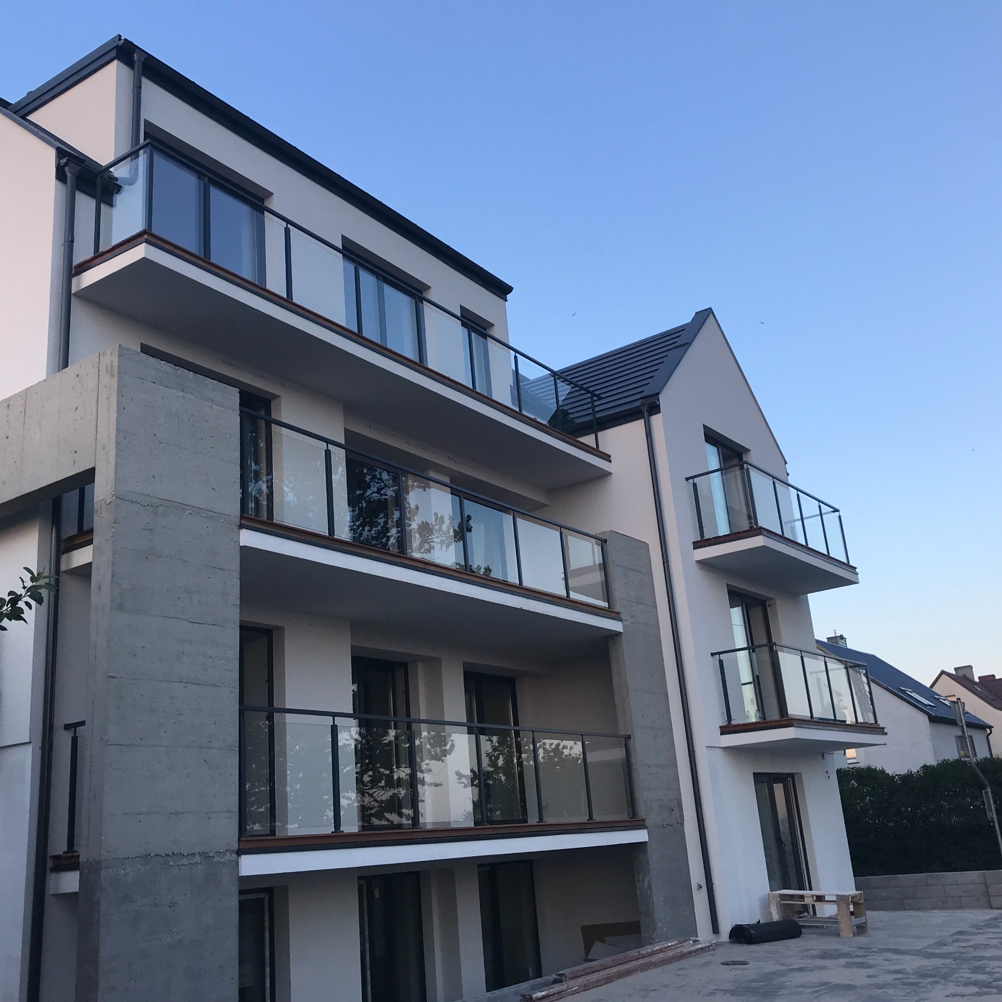 Nowoczesny budynek wielorodzinny z balkonami zabezpieczonymi przezroczystymi panelami ze szkła, osadzonymi w czarnych ramach, widoczne betonowe elementy konstrukcyjne budynku.