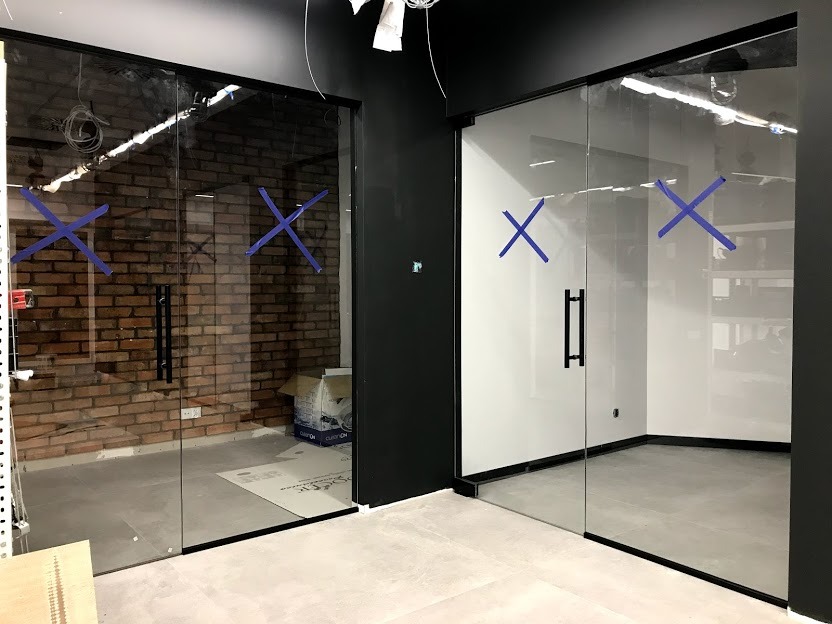 Dwie szklane ścianki działowe z czarnymi ramami i uchwytami, zabezpieczone niebieską taśmą krzyżową, w trakcie montażu; jedna z widokiem na ceglaną ścianę, druga na białe wnętrze.