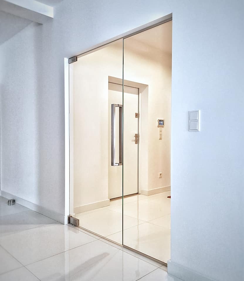 Minimalistyczne, szklane drzwi przesuwne z metalową ramą, prowadzące do jasnego korytarza z białymi płytkami podłogowymi i drzwiami wejściowymi w tle.