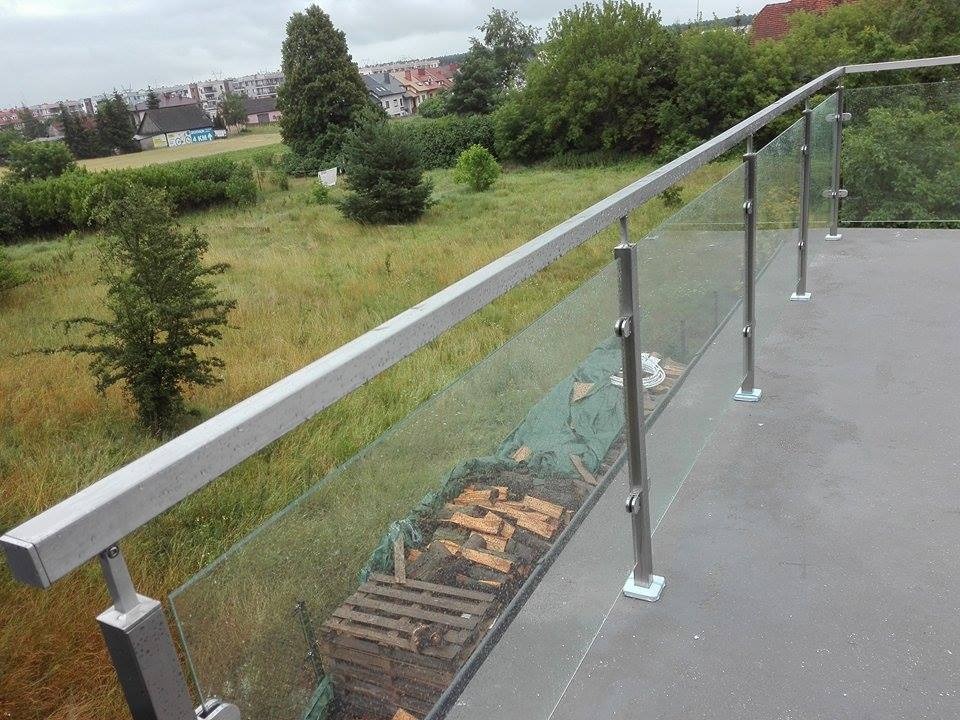 Balkon z balustradą szklaną i metalową poręczą, widok na łąkę i zabudowania miejskie w tle, na podłodze leżą materiały budowlane.