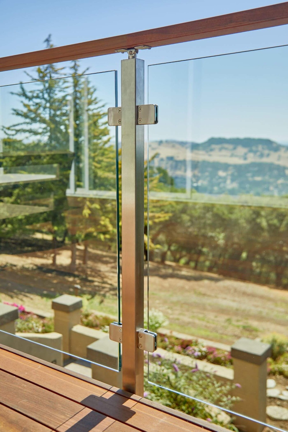 Detale montażu szklanej balustrady na tarasie z drewnianą podłogą i widokiem na wzgórza w tle, z bliska widoczny stalowy słupek mocujący szkło.