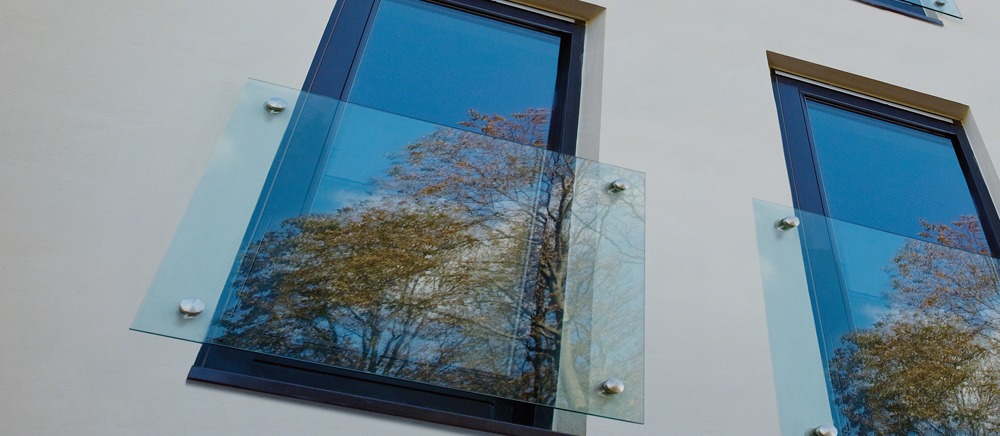 Szklana osłona balkonu z metalowymi mocowaniami przy oknie, odbijająca jesienne drzewo i błękitne niebo.
