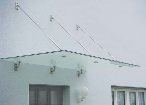 Szklany daszek nad wejściem do budynku, wsparty na metalowych wspornikach i linkach, widoczne okno i oświetlenie zewnętrzne.