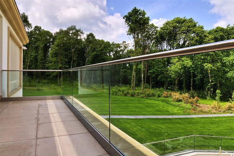 Szklana bariera balkonowa z metalową poręczą, widok na zielony, zadbany ogród i las w tle.