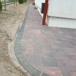 Ułożona, czerwono-szara kostka brukowa obok budynku z czerwoną rurą spustową, widoczne obrzeża i przygotowany teren pod trawnik.