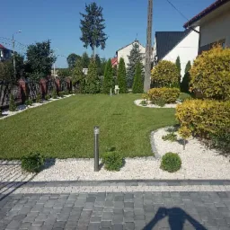 Starannie utrzymany trawnik z równo przyciętą trawą, obrzeża z białego kamienia i ozdobne krzewy w przydomowym ogrodzie, widoczne oświetlenie ogrodowe i fragment kostki brukowej.