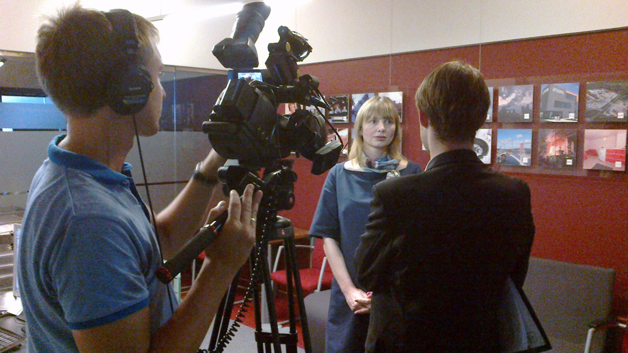 Kobieta w sukience udziela wywiadu przed kamerą w studio z czerwonym tłem i zdjęciami.