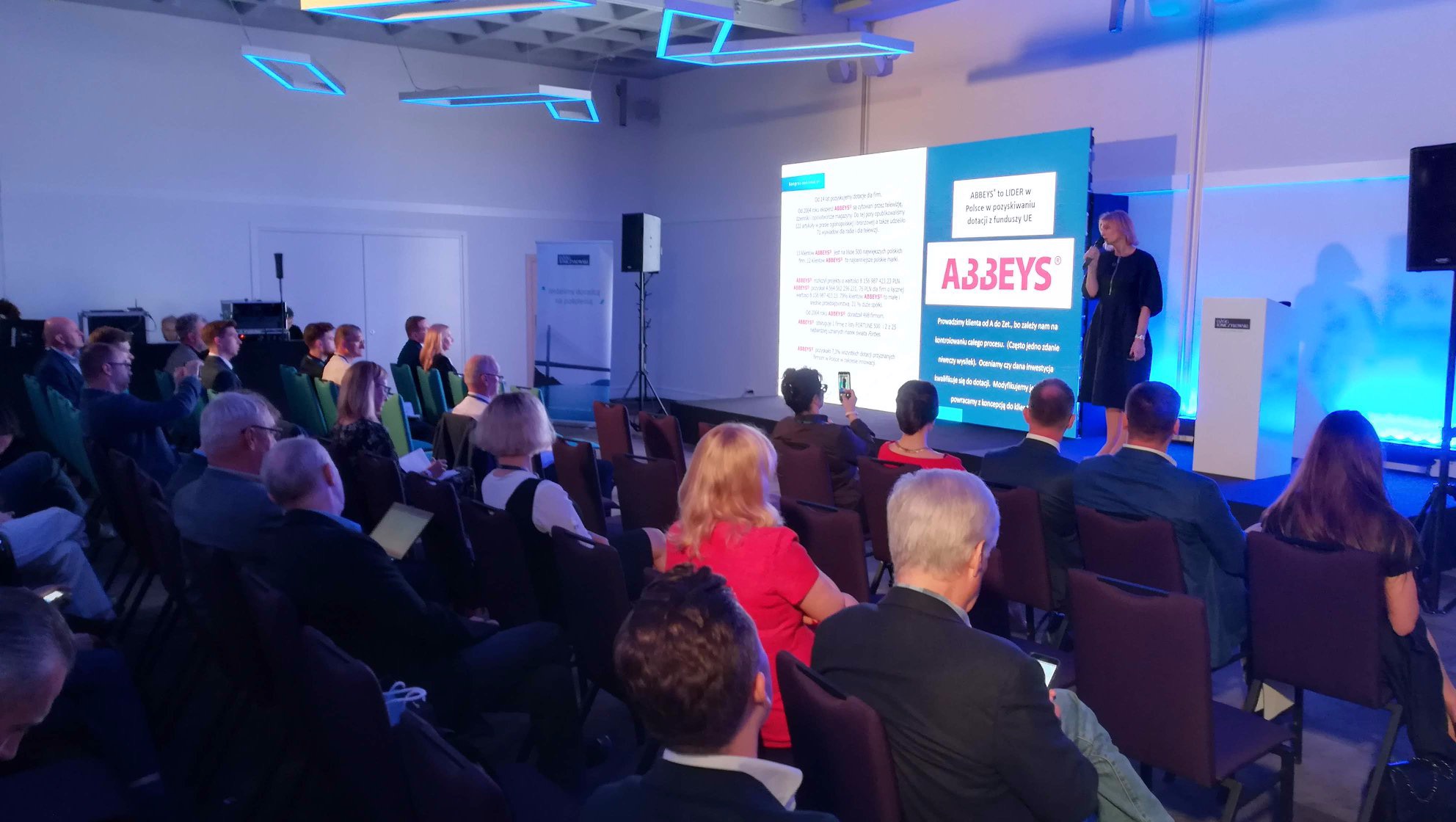 Prezentacja firmy konsultingowej podczas konferencji. Kobieta w czarnej sukience przemawia do zgromadzonych słuchaczy, na ekranie za nią widoczne logo firmy i informacje o pozyskiwaniu funduszy UE.