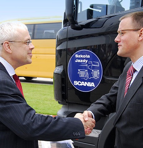 Uścisk dłoni dwóch mężczyzn w garniturach na tle czarnej kabiny ciężarówki Scania z naklejką 'Szkoła Jazdy'. W tle widać żółty autobus.