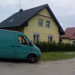 Żółty dom jednorodzinny z ciemną dachówką widziany z boku, z zaparkowanym turkusowym vanem na szutrowym podjeździe.