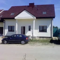 Jasny dom z ciemnym dachem i białymi kolumnami przy wejściu, zaparkowany ciemnoniebieski samochód osobowy przed budynkiem, widok na elewację po remoncie.