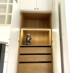 Otwarta szafka kuchenna z trzema drewnianymi szufladami i podświetleniem LED, na półce ekspres do kawy, białe fronty szafek.