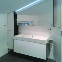 Nowoczesna łazienka z białą, wiszącą szafką pod umywalkę z szufladami, lustrzaną szafką górną z oświetleniem LED i czarną wnęką obok, na ciemnej płytce podłogowej.