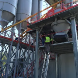 Montaż konstrukcji stalowej z silosami, pracownicy na drabinie i platformie, widoczny fragment ciężarówki w tle.