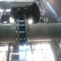 Połączenie kołnierzowe rurociągu przemysłowego z widocznym numerem '130' na rurze, zamontowane wewnątrz hali.