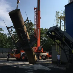 Pomarańczowy dźwig podnosi pionowo ustawiony element przemysłowy w metalowej konstrukcji, widoczni dwaj pracownicy w kaskach ochronnych, w tle budynki przemysłowe i zieleń.