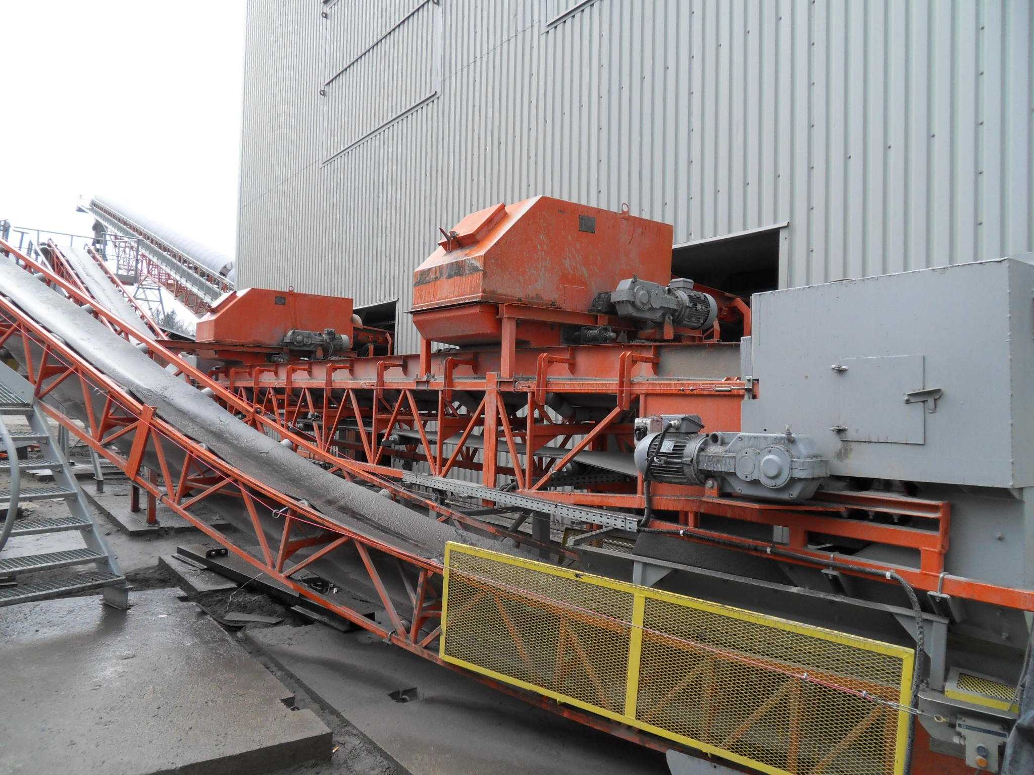 Kompleksowy system przenośników taśmowych w kolorze pomarańczowym, transportujący materiał sypki, z widocznymi silnikami i konstrukcją stalową na tle szarej hali przemysłowej.
