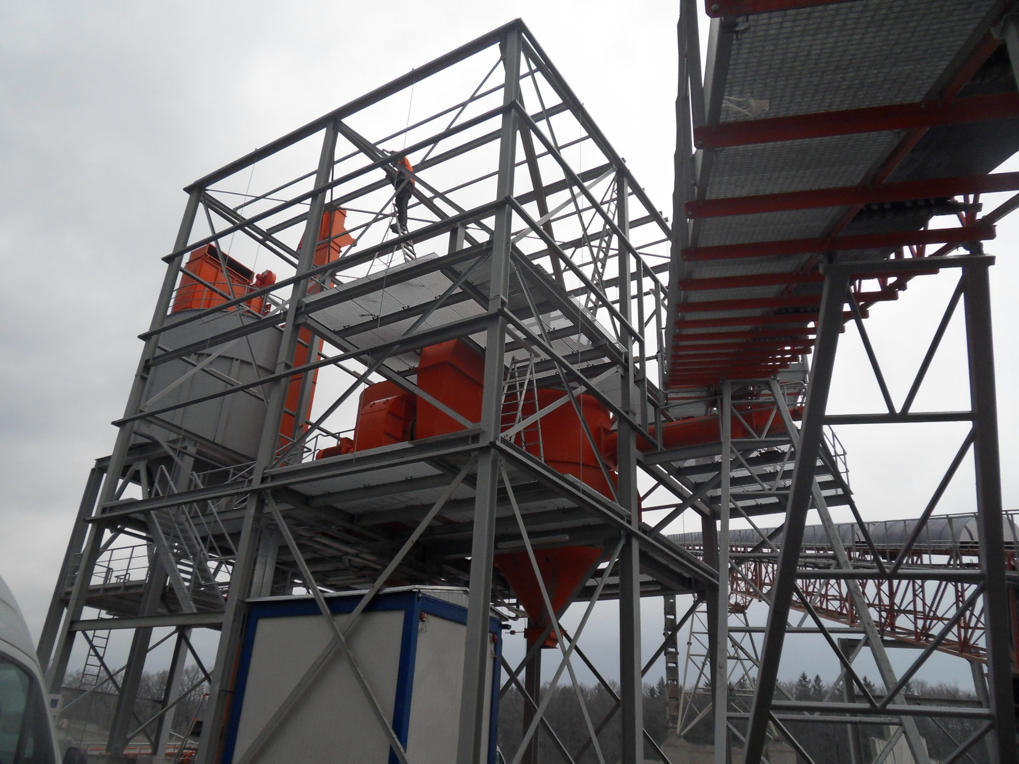 Szkielet budowanej hali stalowej z pomarańczowymi elementami instalacji, widok z dołu na pochmurne niebo, pracownik na drabinie w górnej części konstrukcji.