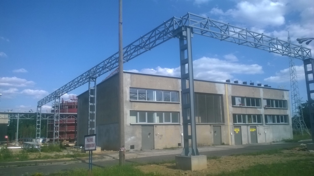 Stalowa konstrukcja hali w trakcie budowy w miejscowości Mirów, widoczna na tle błękitnego nieba z chmurami, z fragmentem istniejącego budynku w tle.