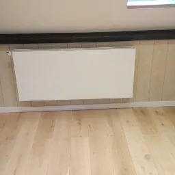 Biały, płaski grzejnik zamontowany na ścianie z boazerią w jasnym odcieniu drewna, widoczny fragment drewnianej podłogi i czarnej belki stropowej.