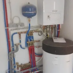 Biały piec gazowy Vaillant z niebieskim zbiornikiem wyrównawczym i białym zasobnikiem ciepłej wody, podłączone rurami miedzianymi i plastikowymi w kolorach niebieskim i czerwonym, na tle białej...