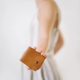 Prezentacja skórzanego portfela w odcieniach brązu, trzymanego w dłoni kobiety w sukience na tle jasnego wnętrza.