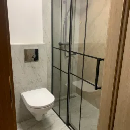 Nowoczesna łazienka z podwieszaną toaletą, marmurowymi płytkami i kabiną prysznicową typu walk-in z czarnymi profilami.