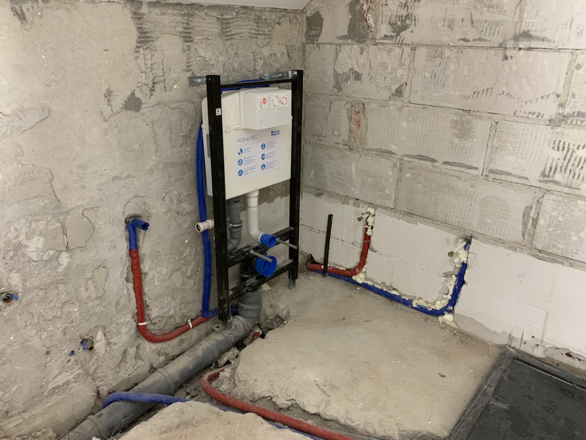 Stelaż podtynkowy WC marki Roca Active WC, z widocznymi niebieskimi i czerwonymi rurami instalacji wodnej w surowym wnętrzu łazienki przed wykończeniem, z częściowo otynkowanymi ścianami.