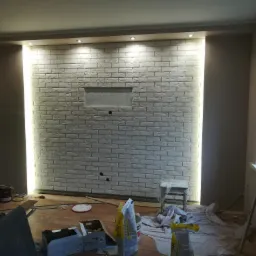 Wnętrze w trakcie wykańczania z białą ceglaną ścianą podświetloną ledami, widoczne narzędzia, wiaderka z farbą i zakryta folią podłoga