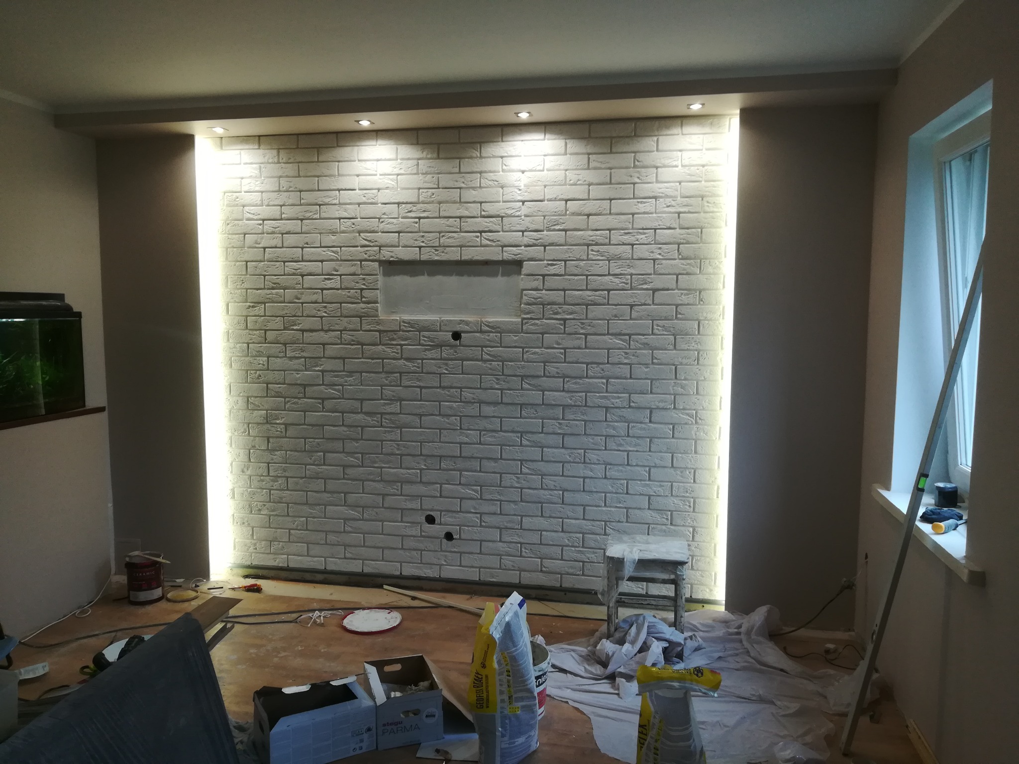 Wnętrze w trakcie wykańczania z białą ceglaną ścianą podświetloną ledami, widoczne narzędzia, wiaderka z farbą i zakryta folią podłoga
