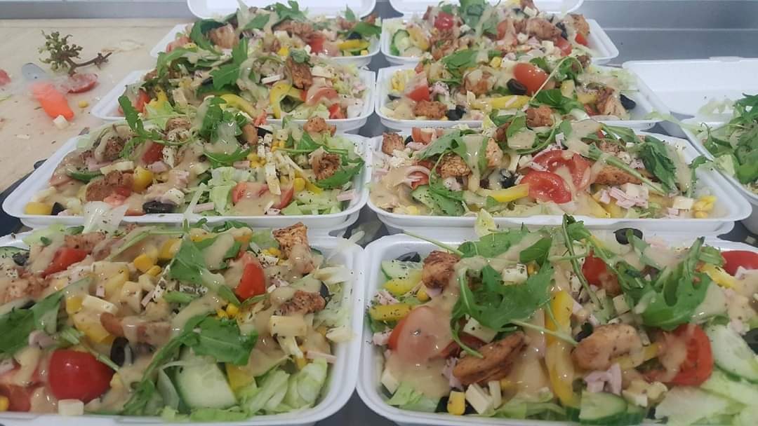 Przygotowane porcje sałatek z grillowanym kurczakiem, pomidorami, ogórkami, kukurydzą, oliwkami i sosem w białych, styropianowych pojemnikach na stole.