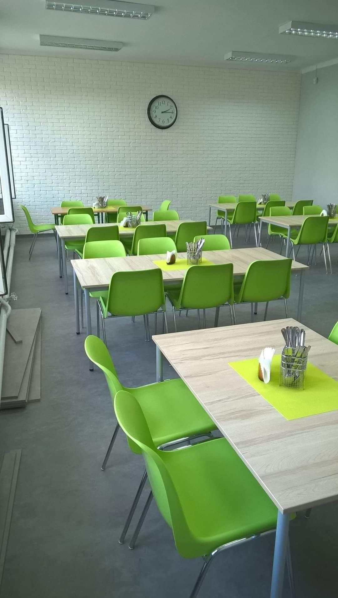 Jasna sala jadalna z białymi ceglanymi ścianami, zegarem i rzędami stołów z jasnozielonymi krzesłami, nakrytych serwetkami i sztućcami, przygotowana do posiłku.