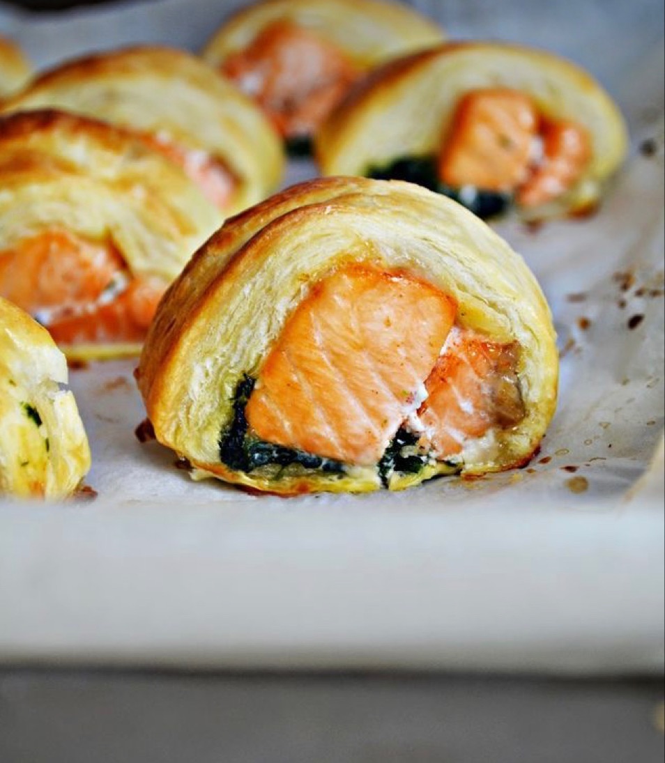 Pojedyncze porcje łososia zapieczonego w cieście francuskim z dodatkiem szpinaku i kremowego sera, ułożone na papierze do pieczenia.