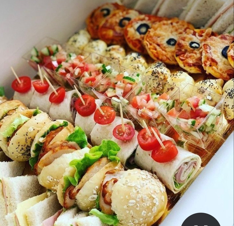 Zestaw przekąsek na tacy, widoczne mini kanapki z kurczakiem i sałatą, tortille z szynką, koreczki z pomidorkami koktajlowymi, sałatki warzywne w małych pojemnikach i mini pizze, przygotowane przez...