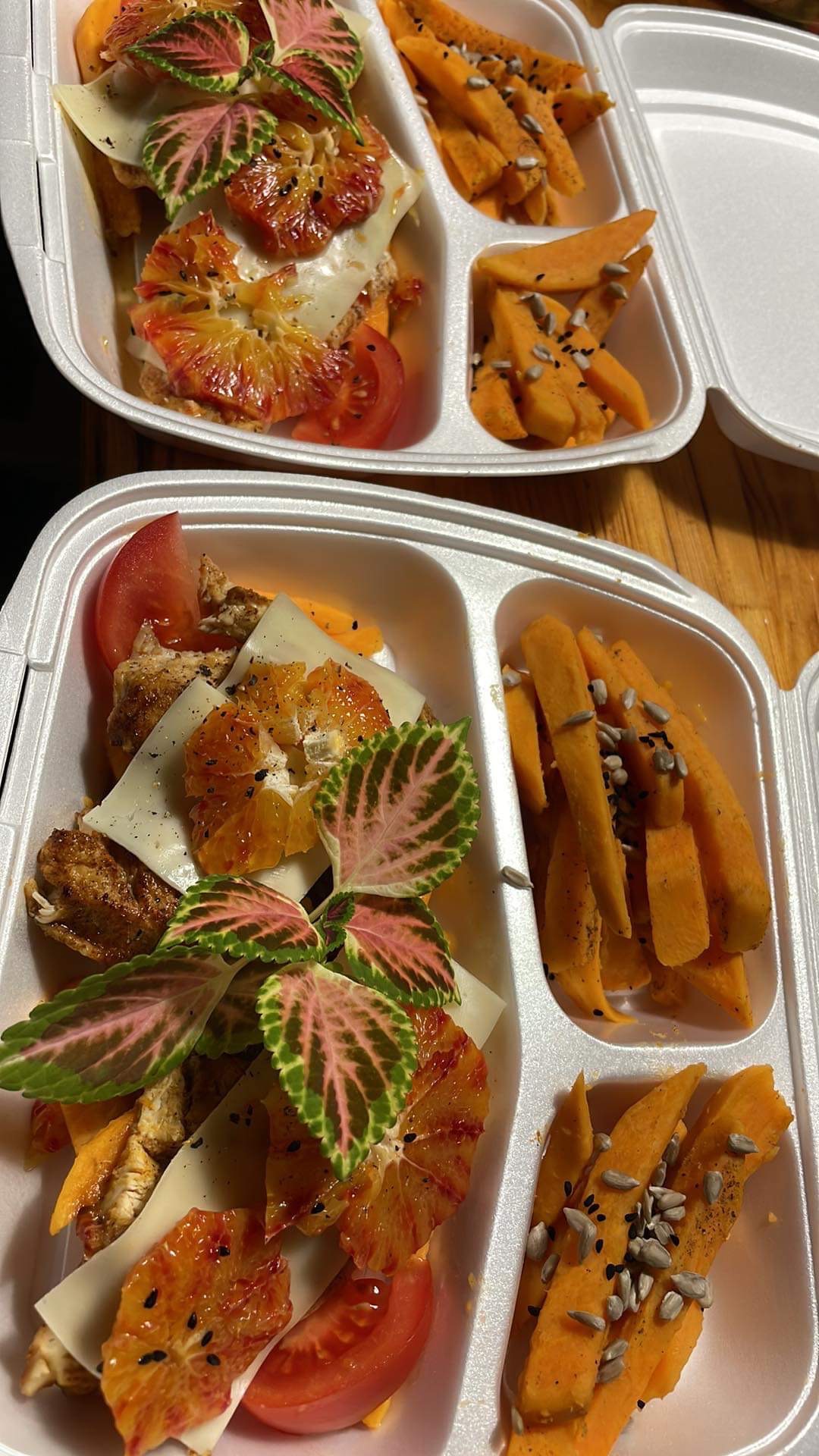 Dwa białe pojemniki z jedzeniem na wynos: kanapka z serem, pomarańczą, pomidorem i dekoracyjnymi liśćmi oraz frytki z batatów posypane ziarnami słonecznika.