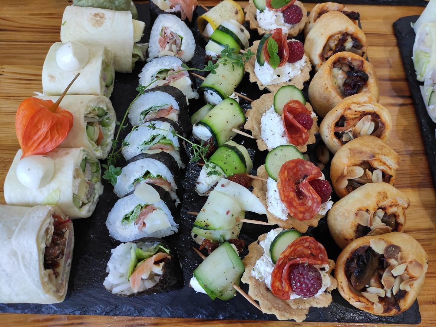 Różnorodny wybór przekąsek na czarnej desce: roladki z tortilli z sosem, sushi z łososiem i warzywami, szaszłyki z cukinii i suszonych pomidorów, tartaletki z kremem, salami i malinami...