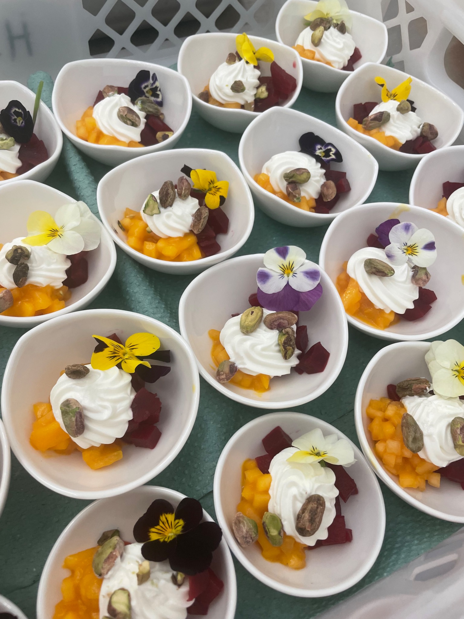Deser w małych białych miseczkach: kostki mango i buraka, bita śmietana, pistacje i jadalne kwiaty fiołka.