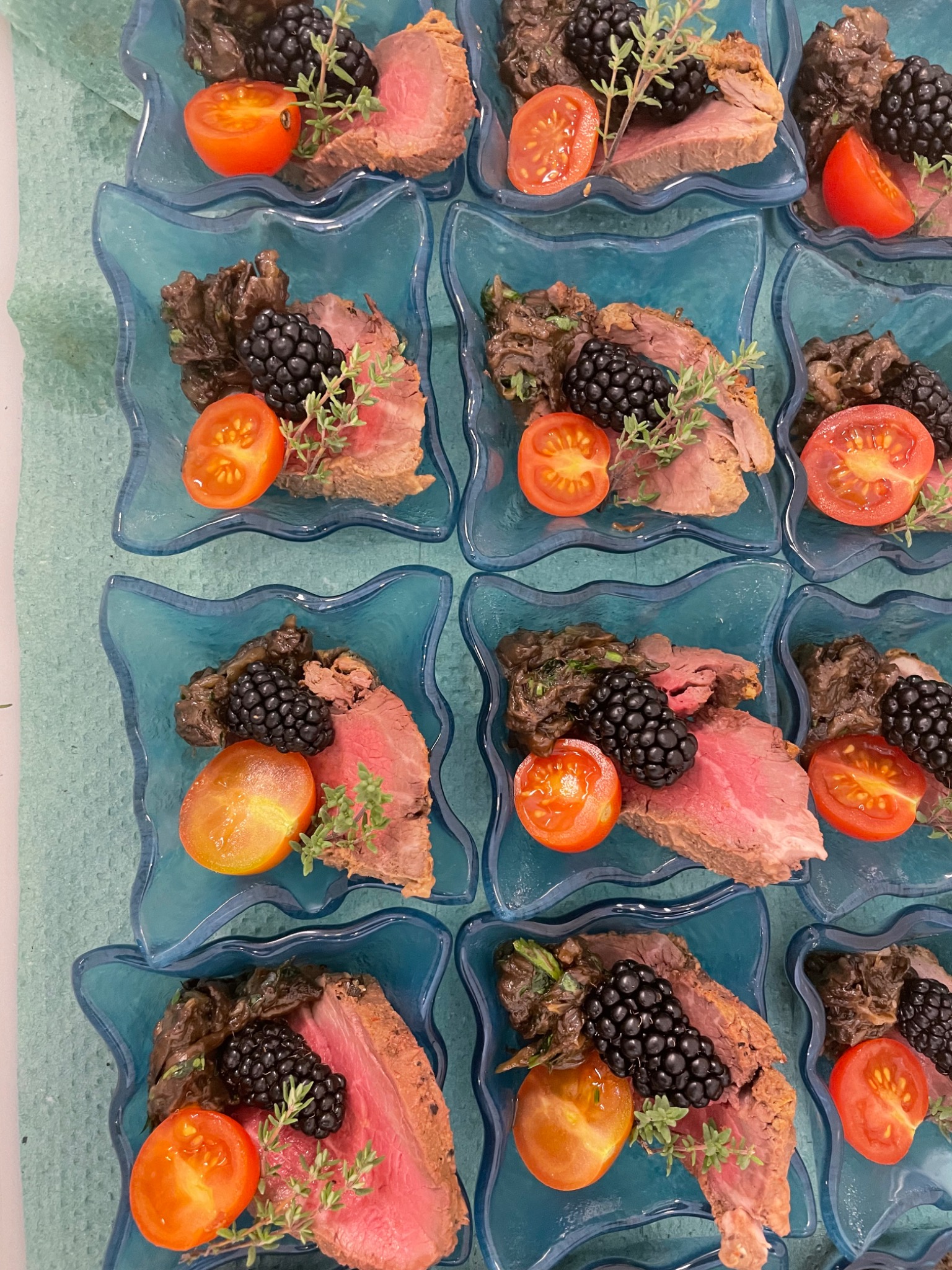 Porcje pieczonej wołowiny z sosem, jeżynami, pomidorkami koktajlowymi i gałązką tymianku, ułożone w niebieskich, dekoracyjnych miseczkach, gotowe do serwowania.