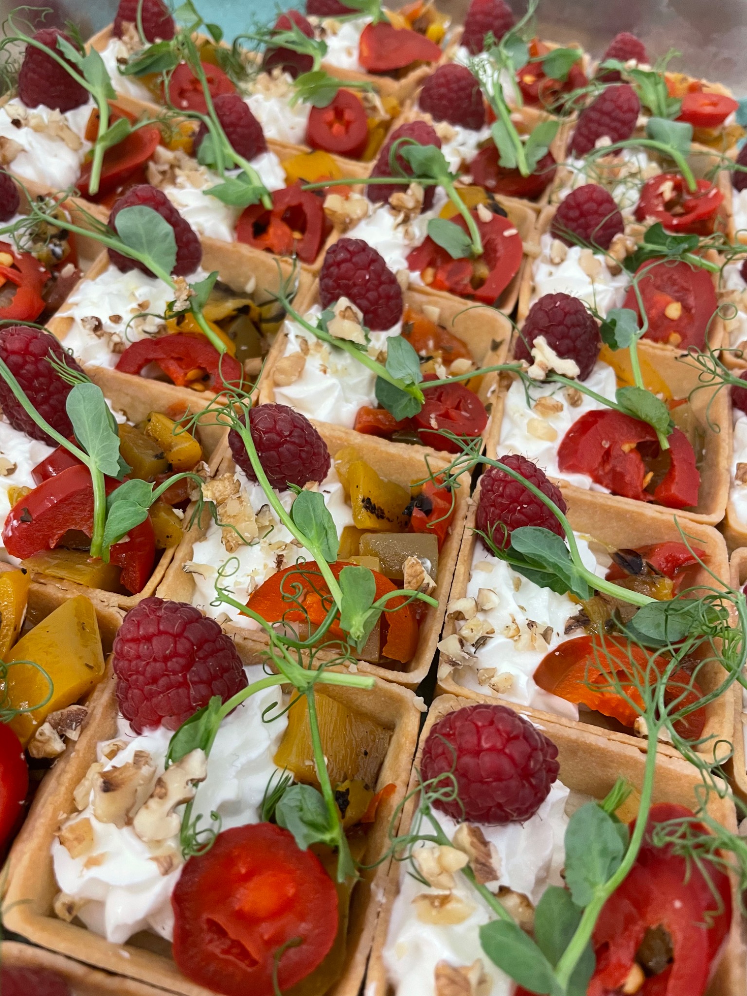 Kwadratowe tartaletki z kremem, grillowaną papryką, malinami, orzechami i pędami groszku ułożone na tacy.