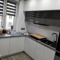 TOP PROJECT Piotr Dul - Nowoczesna kuchnia z białymi szafkami i szarym blatem, czarny zlew i bateria, drewniana deska na płycie. Widok na okno z roletami.